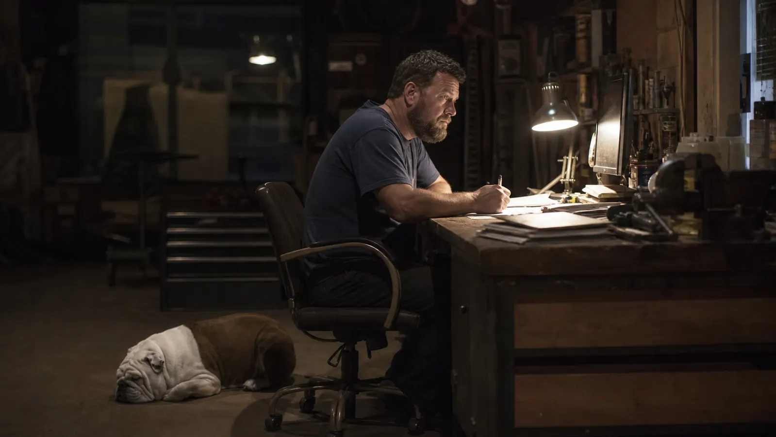 A person reading paperwork at a desk lit by a single desk lamp, the rest of the room in shadow, a bulldog on the floor nearby