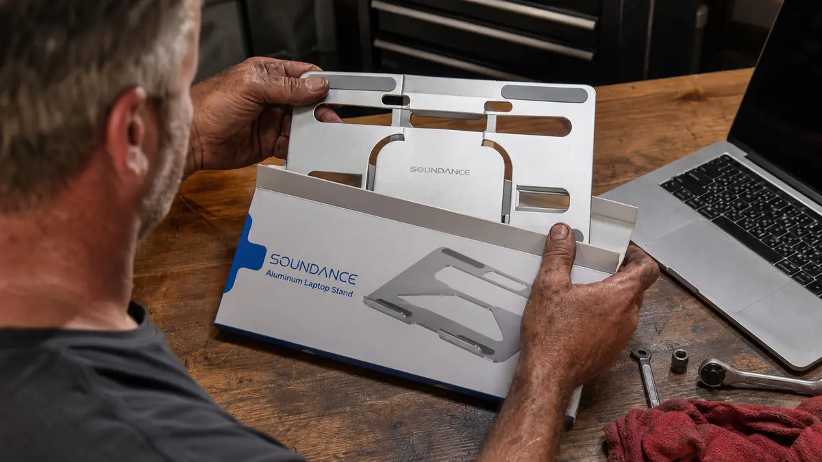 A hand folding a navy blue laptop stand box open on a workbench, the aluminum stand sliding out onto the wood