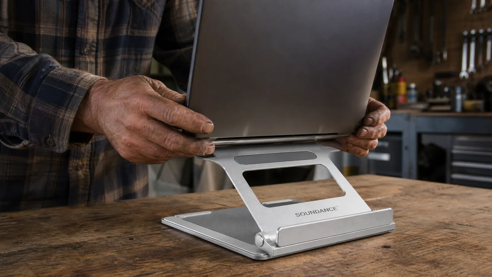 A work-worn hand lowering a laptop onto a SOUNDANCE aluminum stand on a workbench, the folding legs and rubber grip pads clearly visible